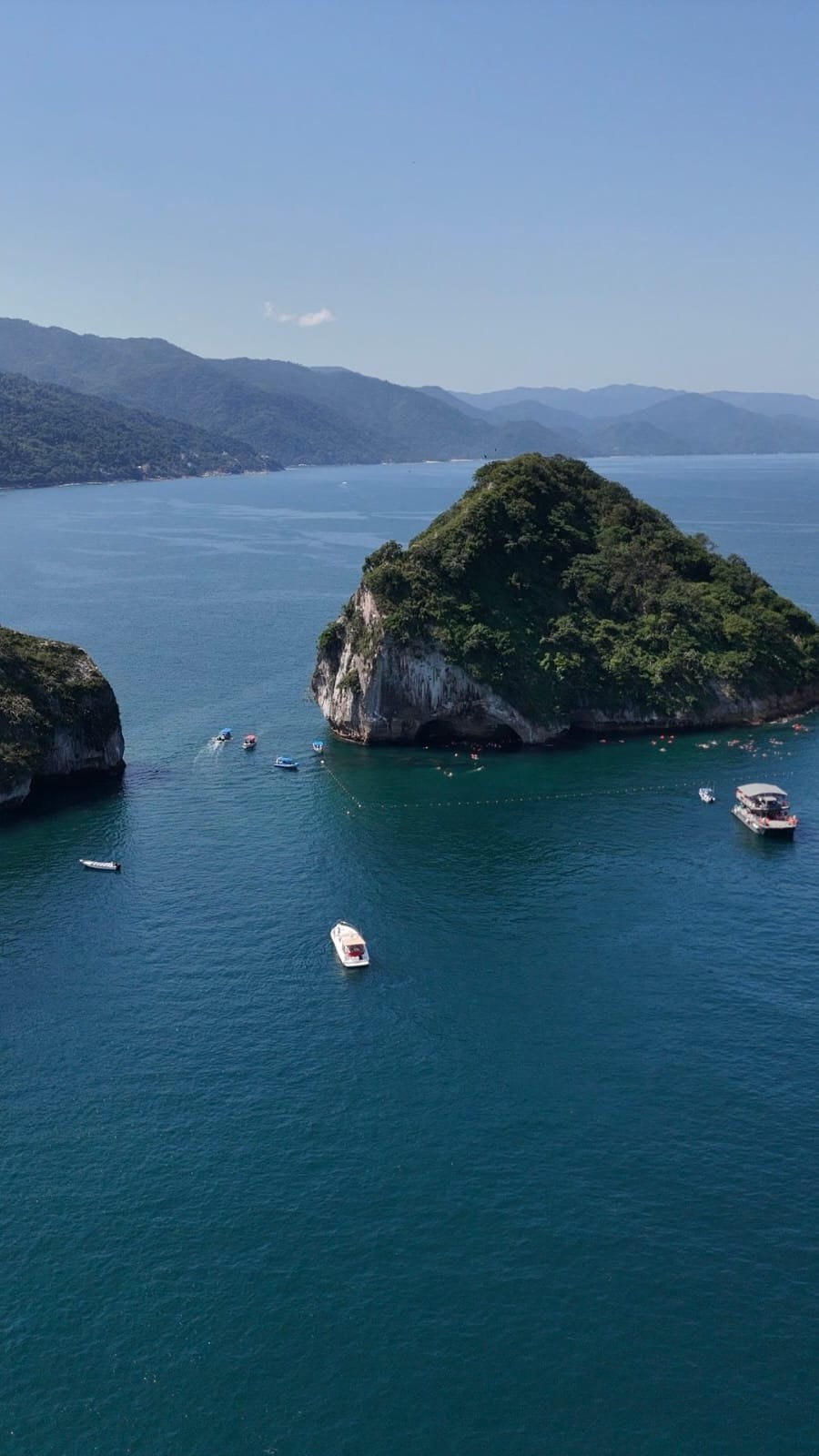 Aerial view of a lush green island with moored boats in turquoise waters, surrounded by mountainous coastline under clear blue sky