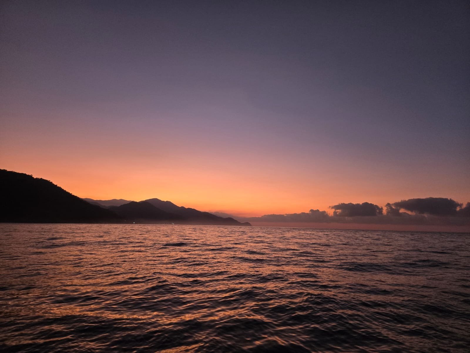 Sunset over calm ocean water with mountainous coastline silhouetted against orange and purple sky