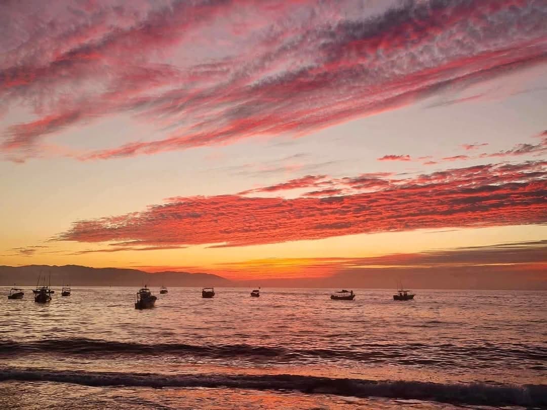 Vivid sunset over calm ocean waters with silhouetted boats moored near shore and dramatic red clouds overhead