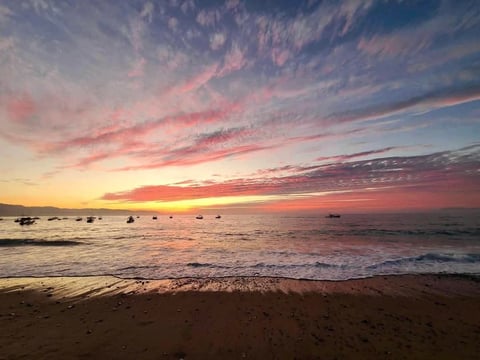 Colorful sunset over a beach with calm waves and scattered boats on the horizon under a pink and orange sky