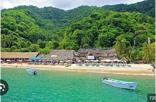Tropical beach resort with thatched-roof bungalows, turquoise water, fishing boats, and lush green mountains in the background