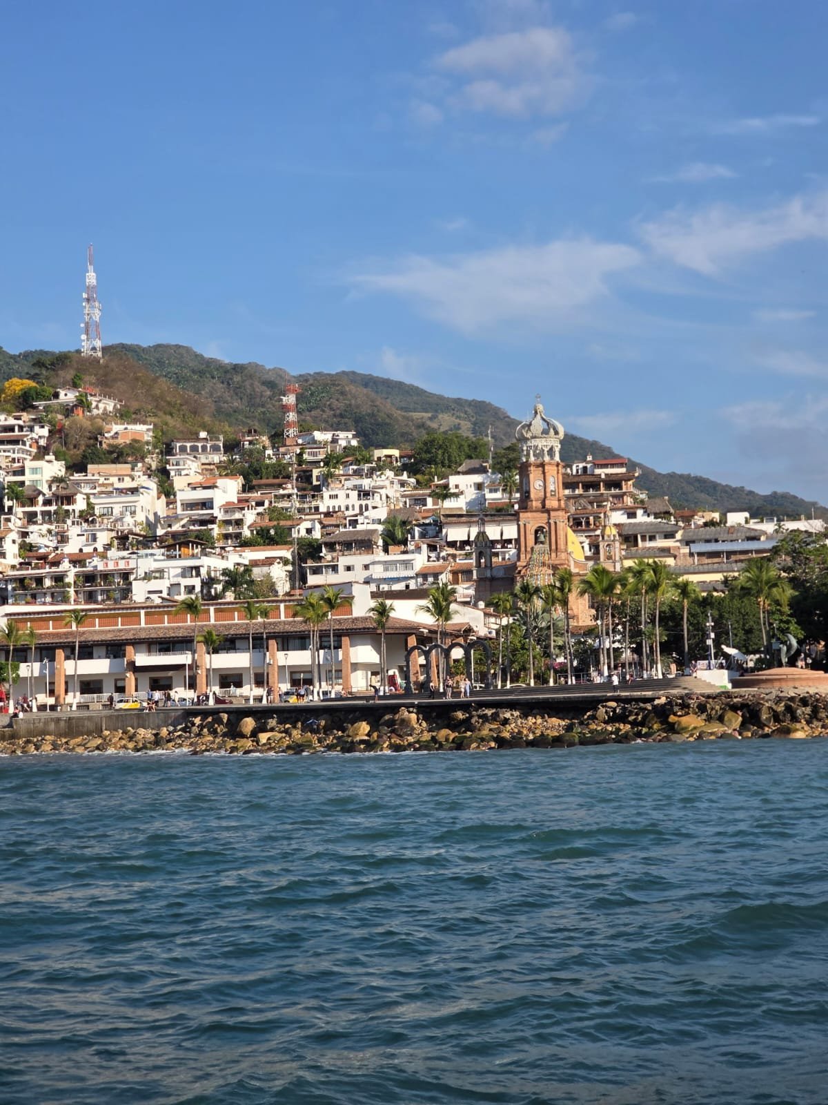 Coastal hillside town with white buildings, a brick church steeple, and a transmission tower viewed from turquoise water